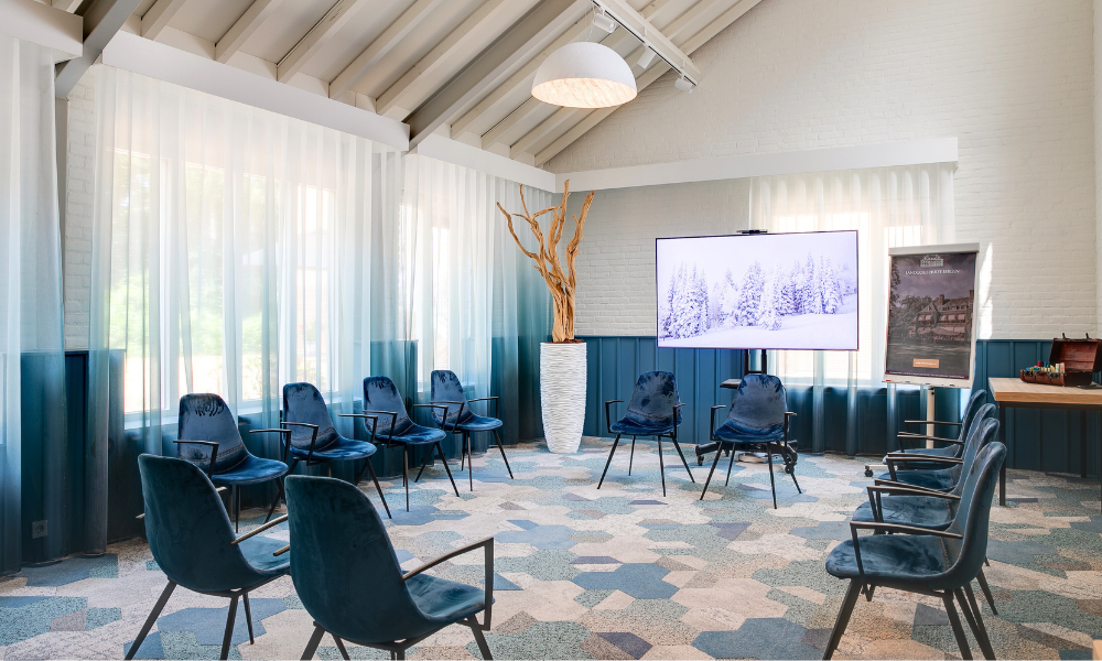 Een goed verlichte vergaderruimte met een cirkel van blauwgroene stoelen, een groot scherm en een presentatiebord. Een decoratieve boom staat in een grote witte pot.