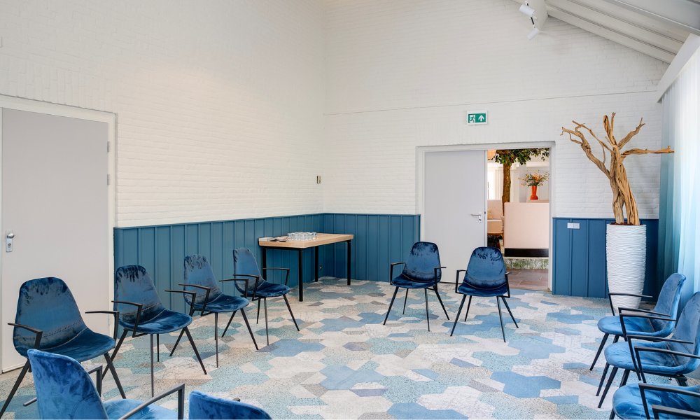 Een lichte vergaderruimte met blauwe fluwelen stoelen in een cirkel. Witte muren en blauwe lambrisering met een vloer met patroon.