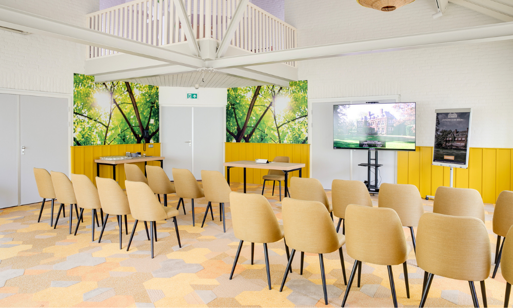 Een moderne, lichte vergaderruimte met rijen bruine stoelen tegenover een groot scherm. Gele accenten en natuurthema-muurkunst zorgen voor een lichte sfeer.