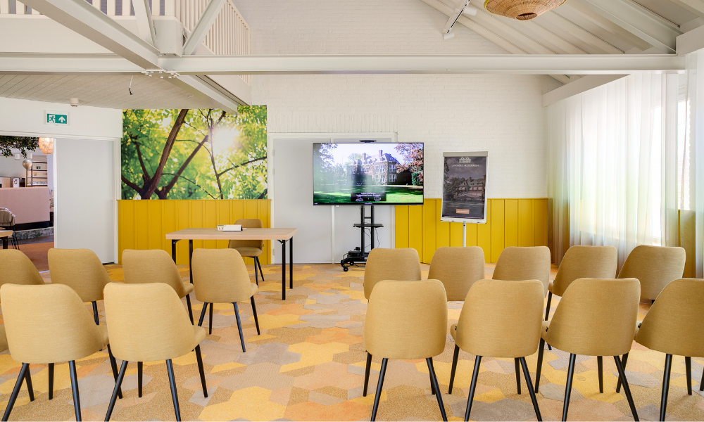 Een lichte vergaderruimte met gele stoelen, een tafel en een groot scherm, met een muurkunstwerk met natuurthema.