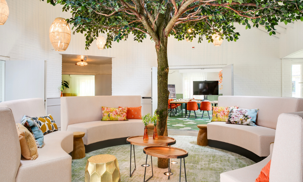 Een moderne lounge met een boom in het midden, omringd door gebogen roze banken, kleurrijke kussens en kleine tafeltjes.