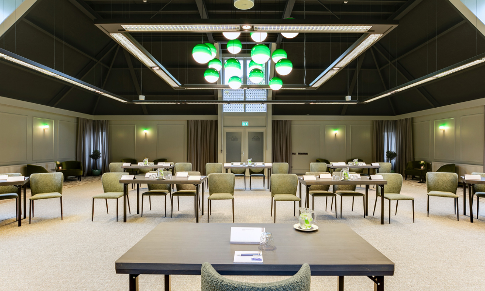 Vergaderruimte met tafels die klaarstaan voor een vergadering. Op de voorgrond staat een grote vierkante tafel en aan het plafond hangen groene bollen.