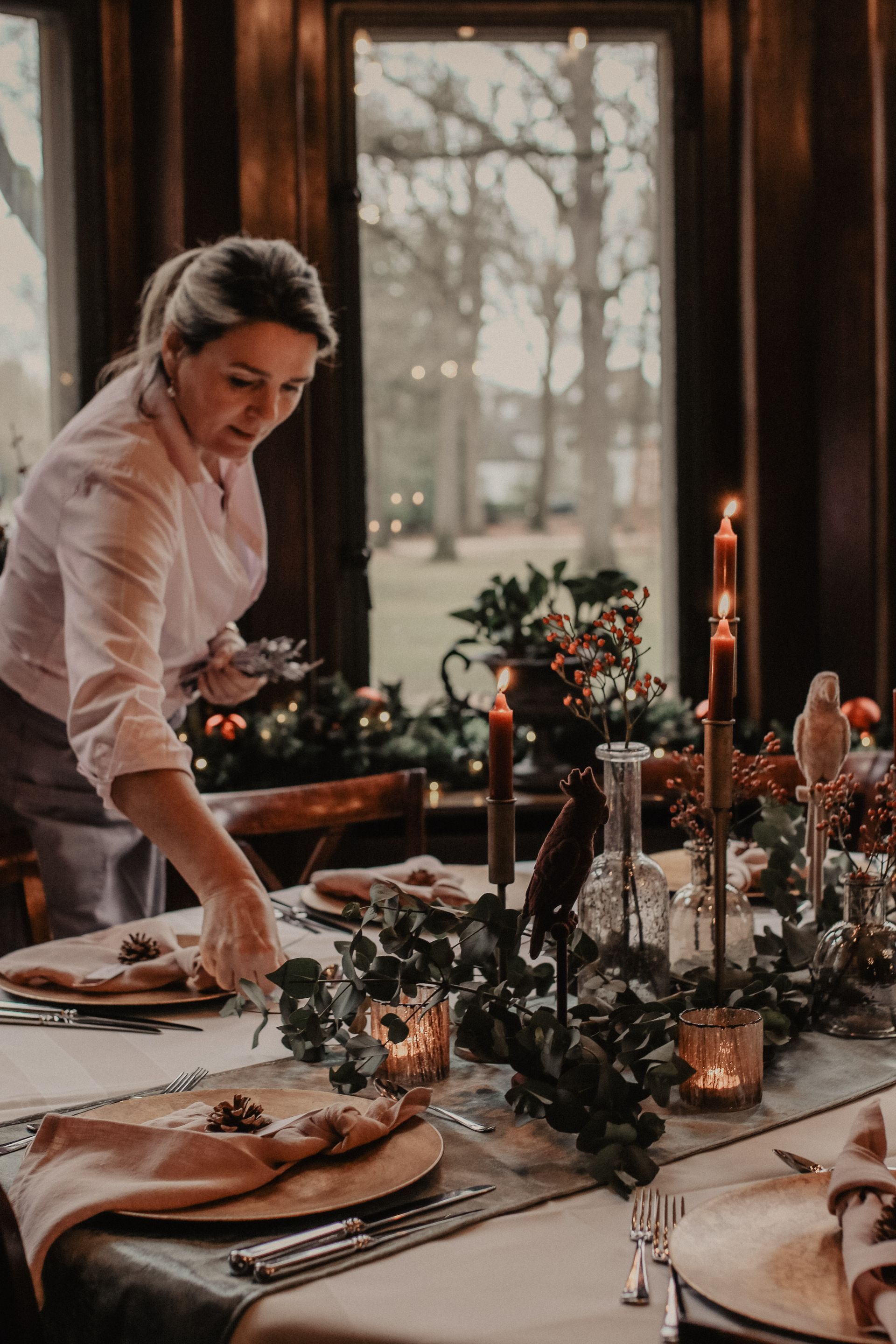 Een vrouw dekt een feestelijke tafel met kaarsen, groen en uitzicht op een landschap door een raam. Warme tinten, gezellig interieur.