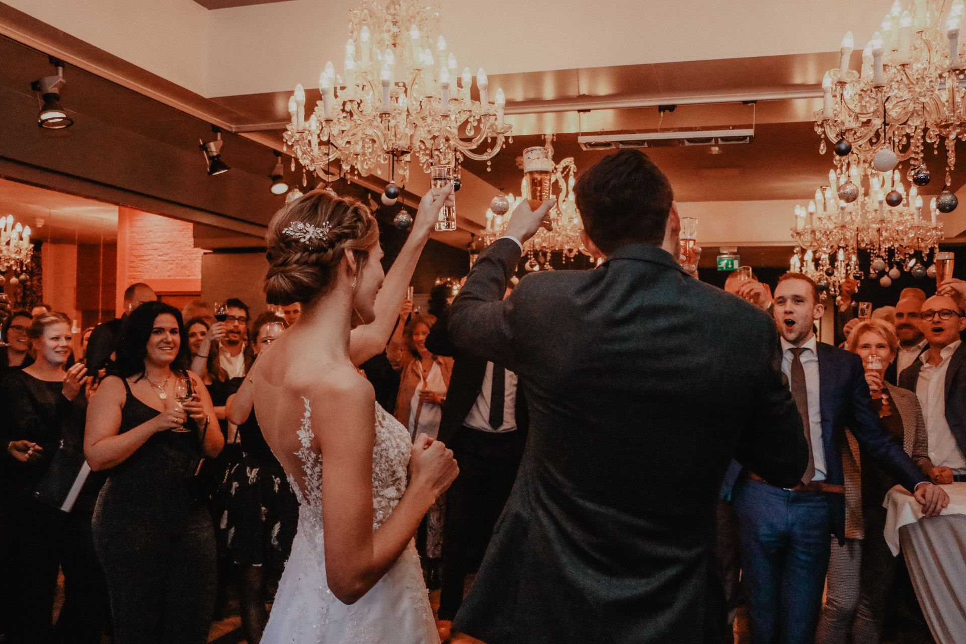 Bruid en bruidegom dansen op hun huwelijksreceptie, omringd door juichende gasten onder kroonluchters.