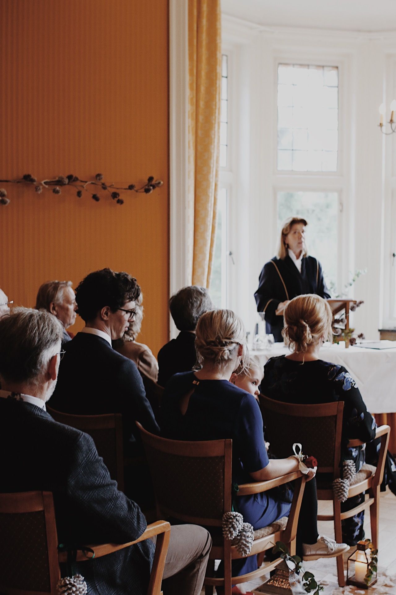 Een persoon in een toga spreekt achter een katheder en richt zich tot een publiek dat op houten stoelen zit in een lichte ruimte.