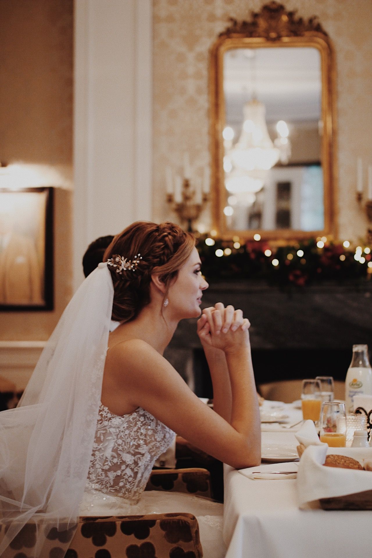 Bruid op een huwelijksreceptie, met haar kin op haar handen. Ze draagt een witte jurk en sluier en zit aan een versierde tafel.