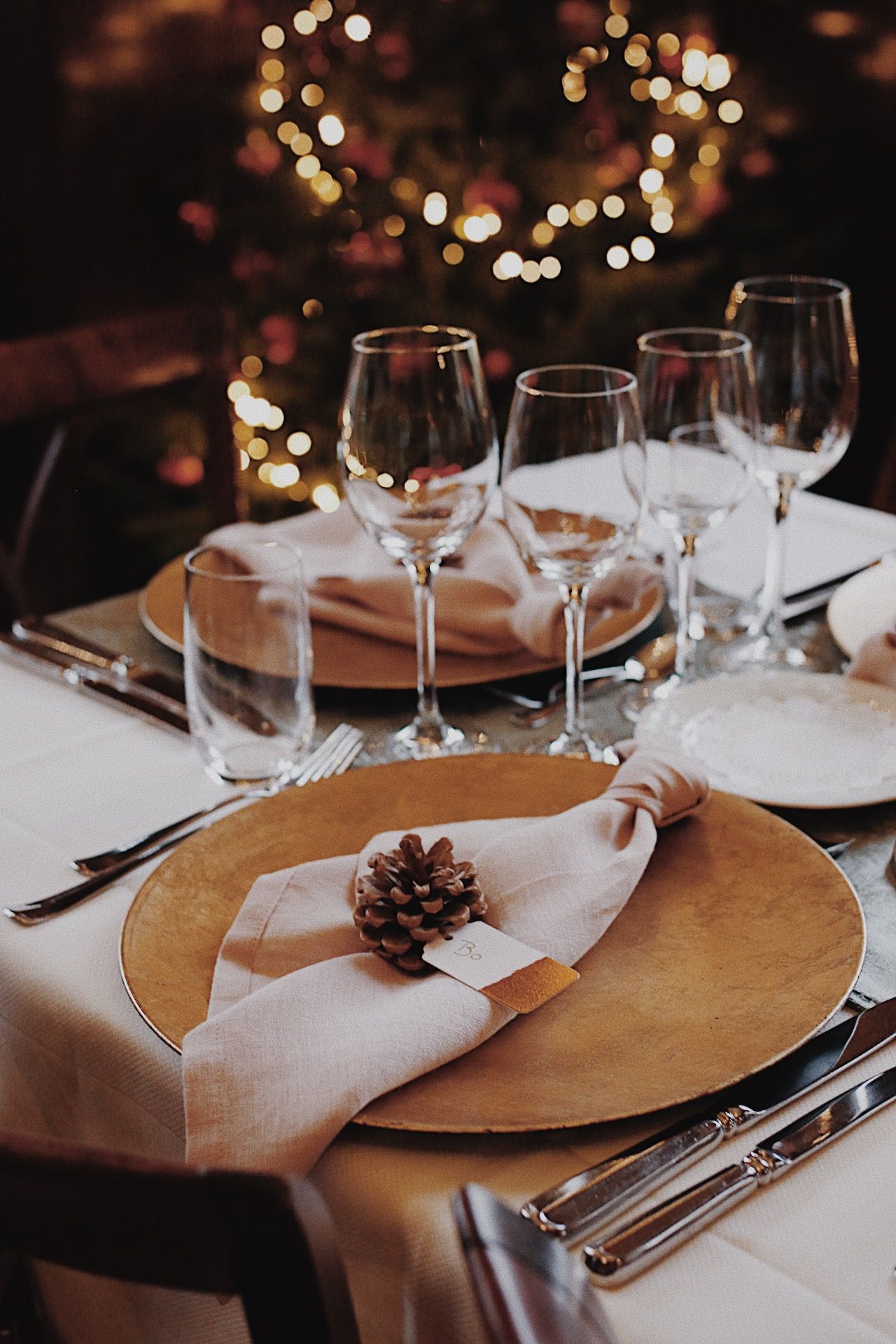 Elegante tafeldekking voor een feestmaaltijd, met gouden onderborden, wijnglazen en een servet versierd met een dennenappel.