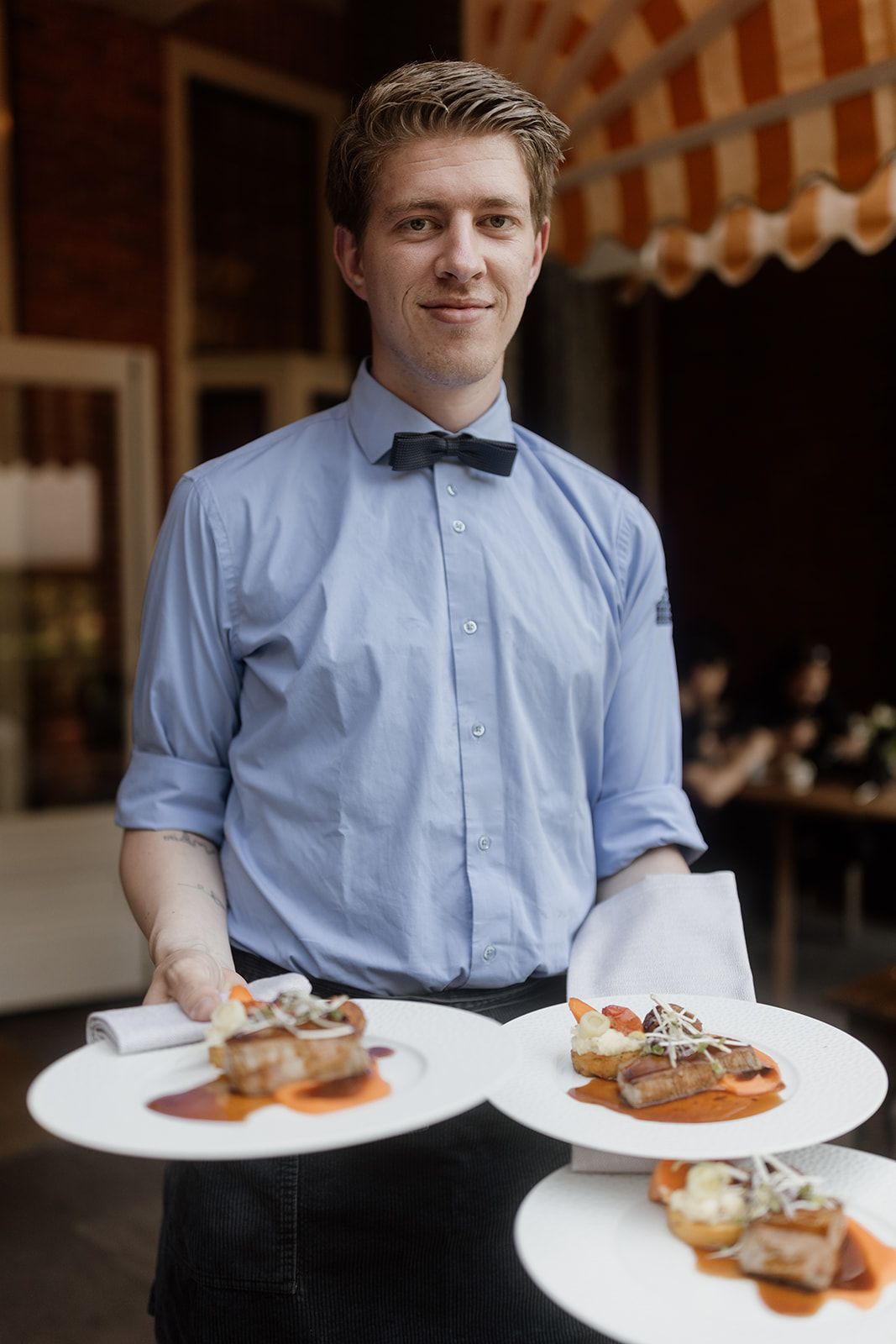 Een ober in een blauw overhemd en vlinderdas lacht terwijl hij drie borden met eten draagt in een openluchtrestaurant.