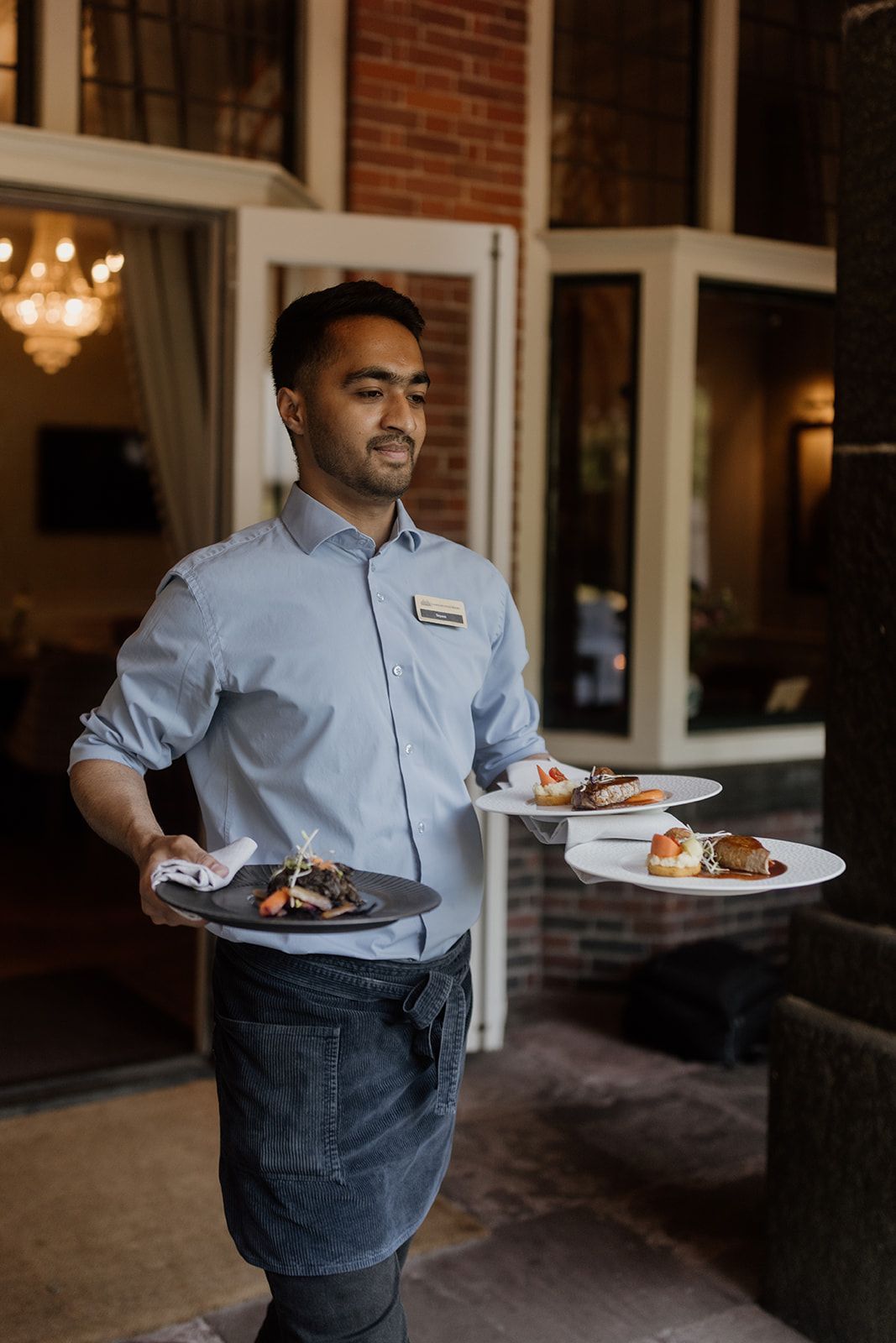 Een glimlachende ober, gekleed in een grijs shirt en schort, draagt borden met eten buiten een restaurant.