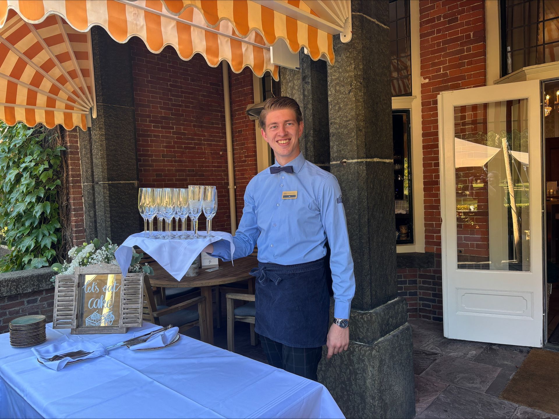 Een glimlachende ober houdt een dienblad met champagneglazen vast buiten een restaurant. Hij staat aan een tafel met champagneglazen en andere uitgestalde items onder een gestreepte luifel.