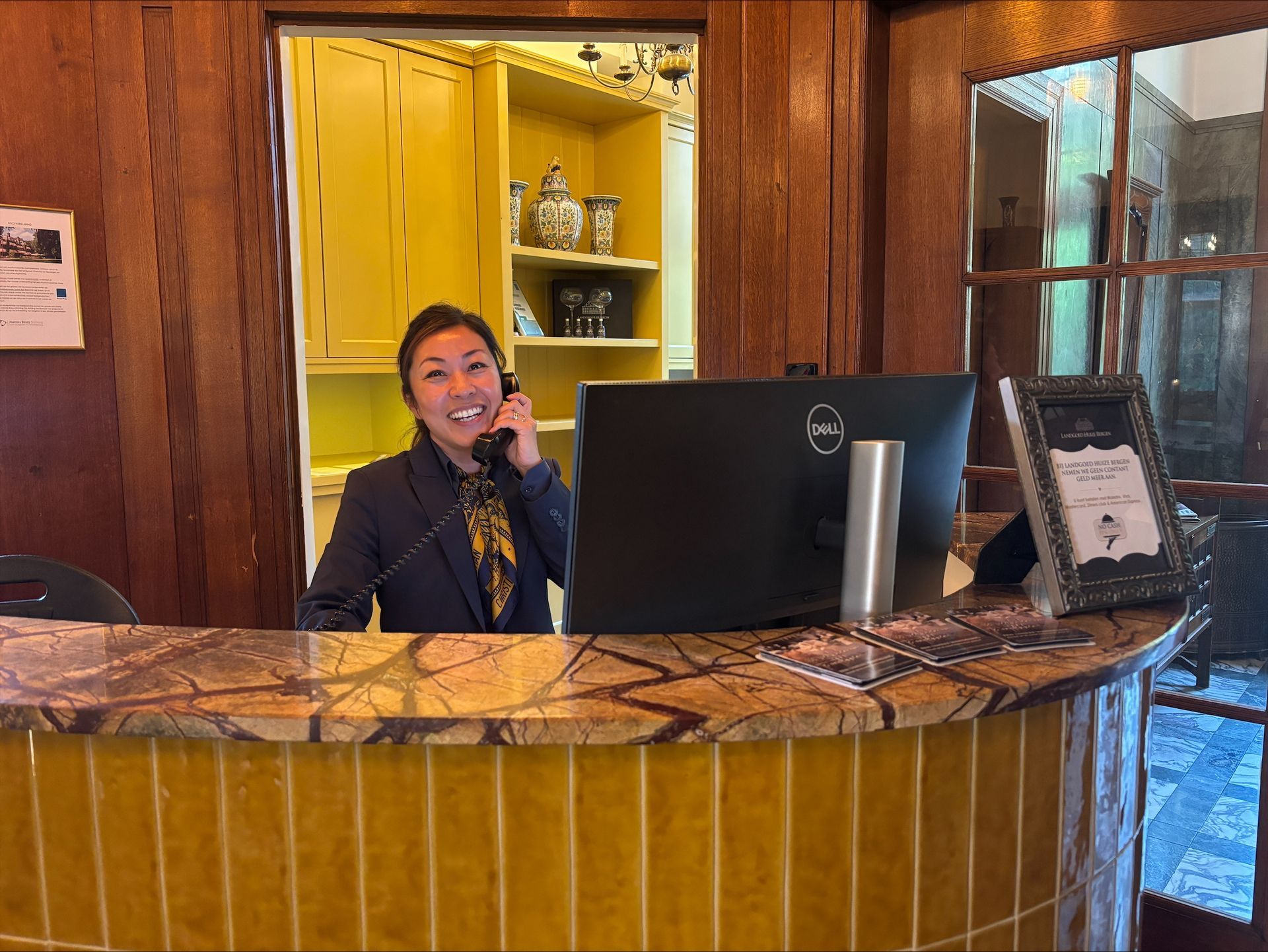Een vrouw aan de receptie, telefonerend. Ze glimlacht, in pak, achter een marmeren balie met een Dell-monitor.