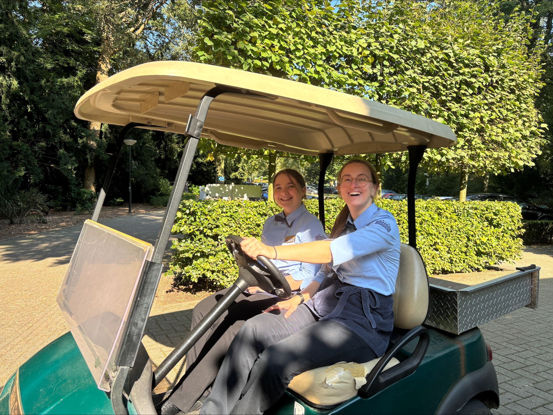 Twee glimlachende vrouwen in lichtblauwe uniformen rijden buiten in een groen golfkarretje. Weelderig groen op de achtergrond.