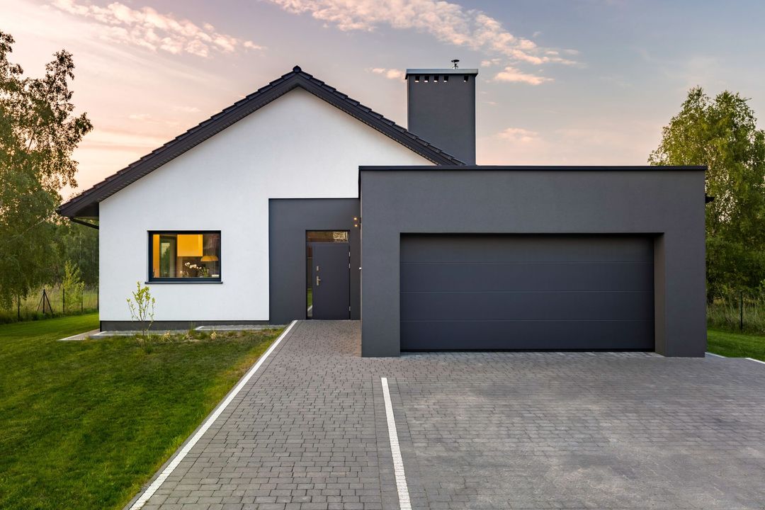 Maison moderne blanche avec garage gris et allée. Cheminée et arbres en arrière-plan.