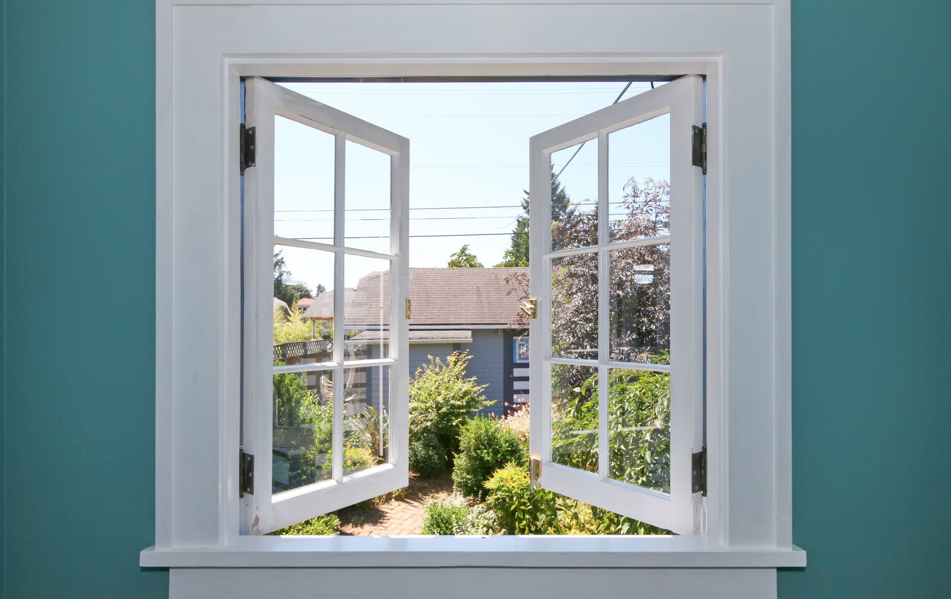 Fenêtre ouverte à cadre blanc donnant sur une cour verdoyante et une maison au toit brun.