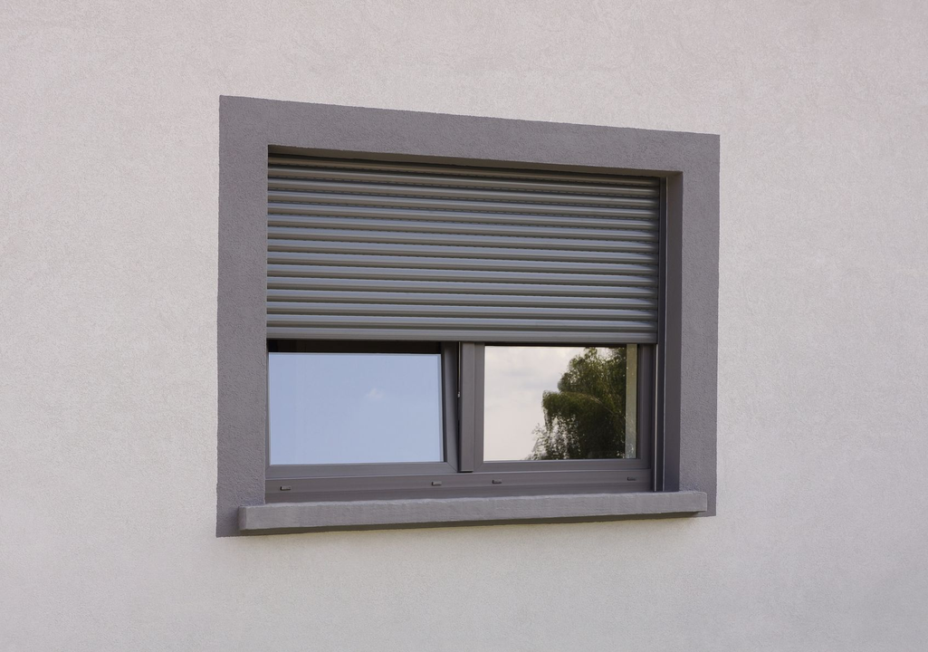 Fenêtre avec volet roulant gris, encadrée de pierre grise sur un mur couleur crème.