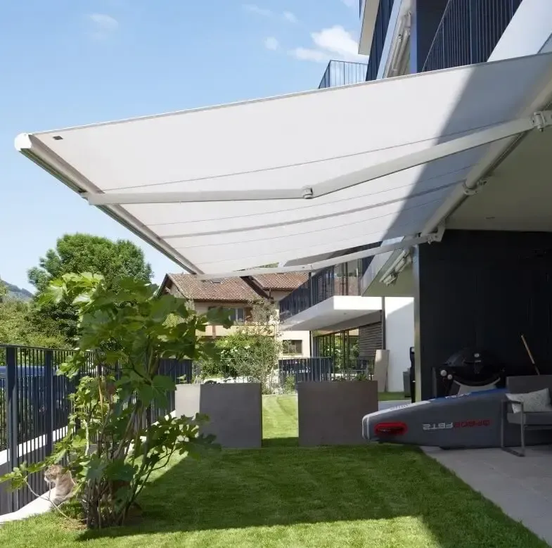 Un store rétractable blanc protège une terrasse avec des plantes en pot et un barbecue.