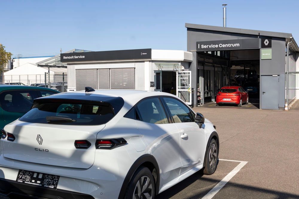Ein weißes Auto parkt vor einem Autohaus, in dessen Werkstatt ein rotes Auto steht.