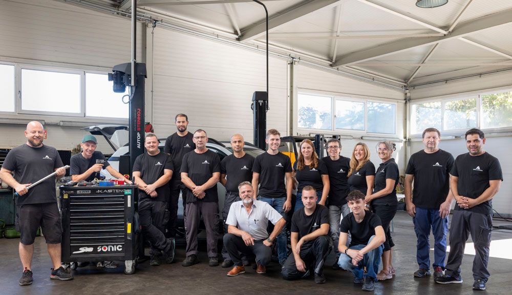 Eine Gruppe von Mechanikern posiert in einer Werkstatt mit einer Hebebühne und einem Werkzeugkasten, einige halten Werkzeuge in den Händen.