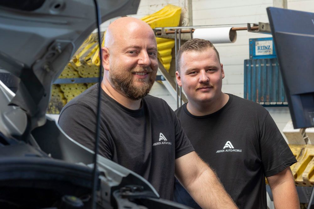 Zwei Mechaniker in schwarzen Hemden lächeln neben einer offenen Motorhaube in einer Werkstatt.