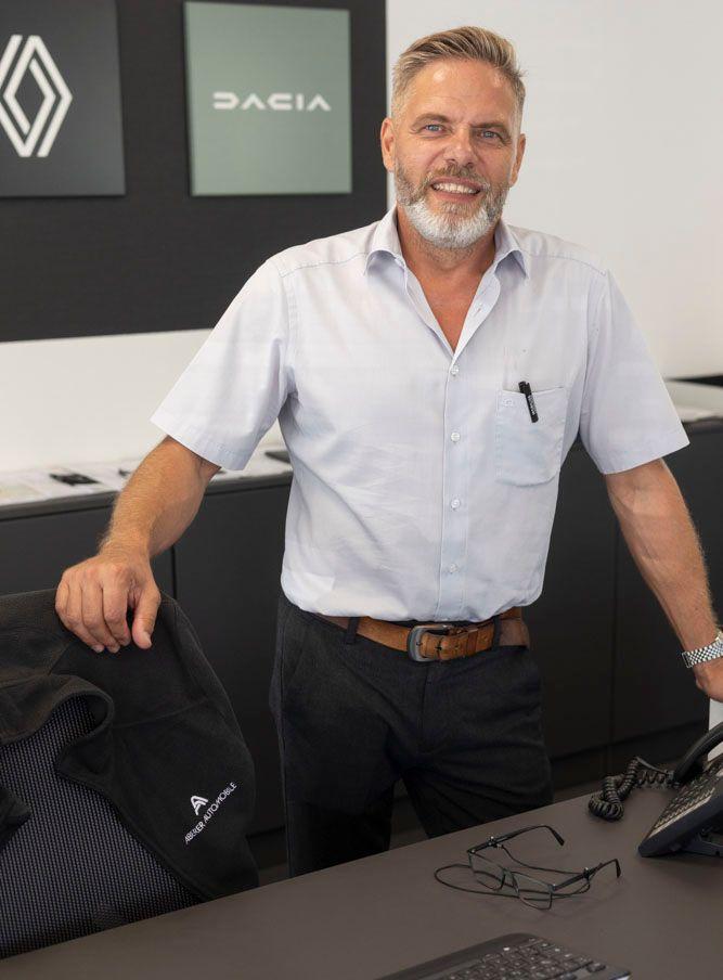 Ein Mann lächelt neben einem Schreibtisch in einem modernen Büro; er trägt ein hellblaues Hemd, im Hintergrund ist das DACIA-Logo zu sehen.