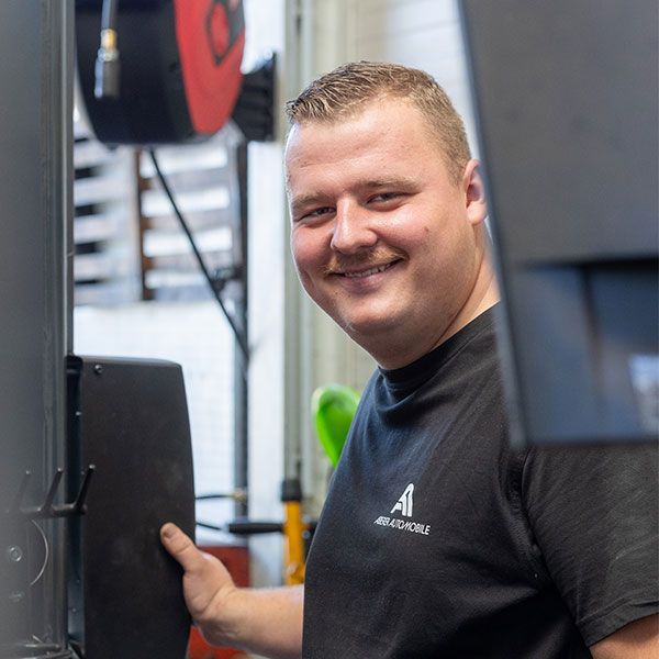 Ein lächelnder Mann in schwarzem Hemd steht neben Geräten in einer Werkstatt und hält ein Bedienfeld in der Hand.