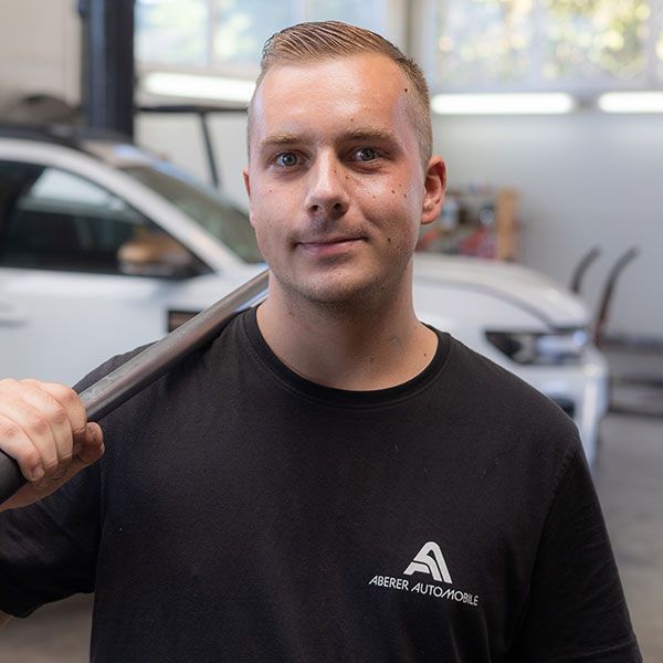 Ein Mann in schwarzem Hemd steht in einer Garage, hält einen Schultergurt und blickt in die Kamera.