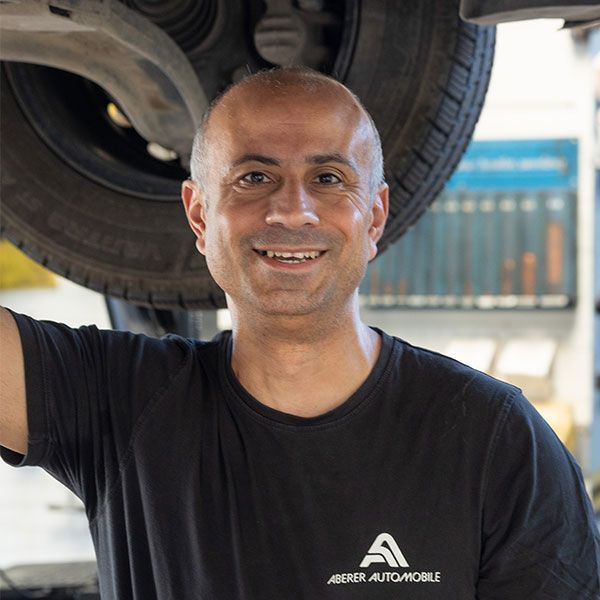 Ein lächelnder Mechaniker steht unter einem angehobenen Auto und trägt ein schwarzes Hemd in einer Autowerkstatt.