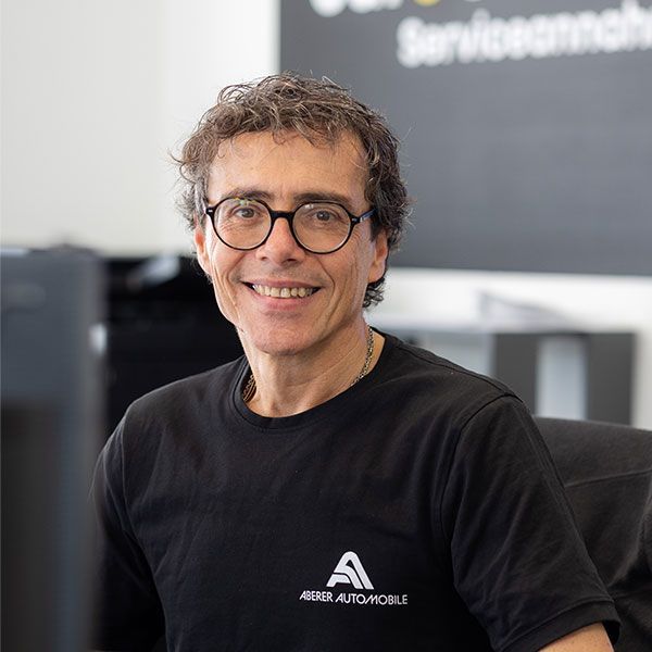 Eine lächelnde Person mit Brille und schwarzem T-Shirt sitzt in einem modernen Büro.