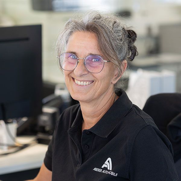 Eine lächelnde Person mit Brille und schwarzem Poloshirt sitzt an einem Schreibtisch mit Computer.