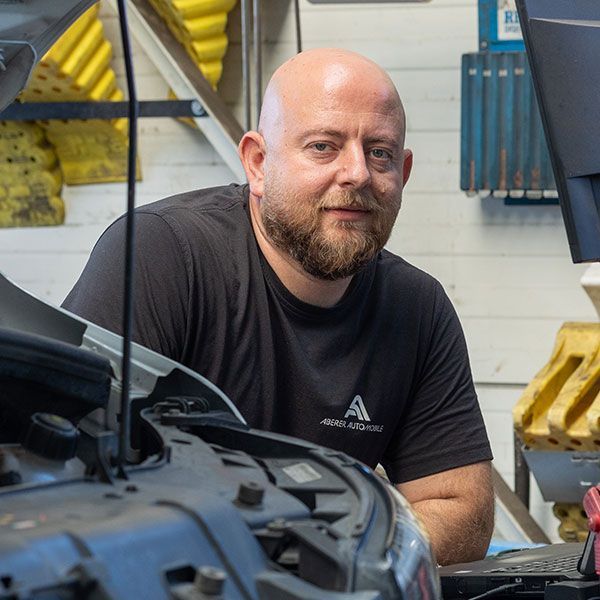 Ein Mechaniker steht in einer Werkstatt neben einer Motorhaube und blickt in die Kamera; hinter ihm befinden sich Regale mit Werkzeugen.