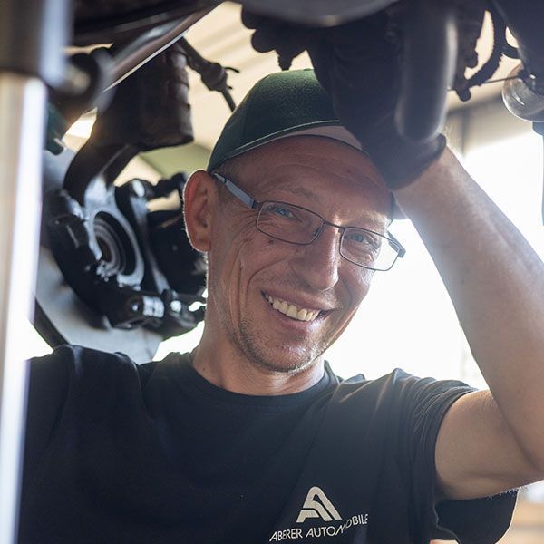Ein lächelnder Mann mit Brille und Kappe arbeitet unter einem Fahrzeug, den Arm in der Nähe einer Maschine erhoben.