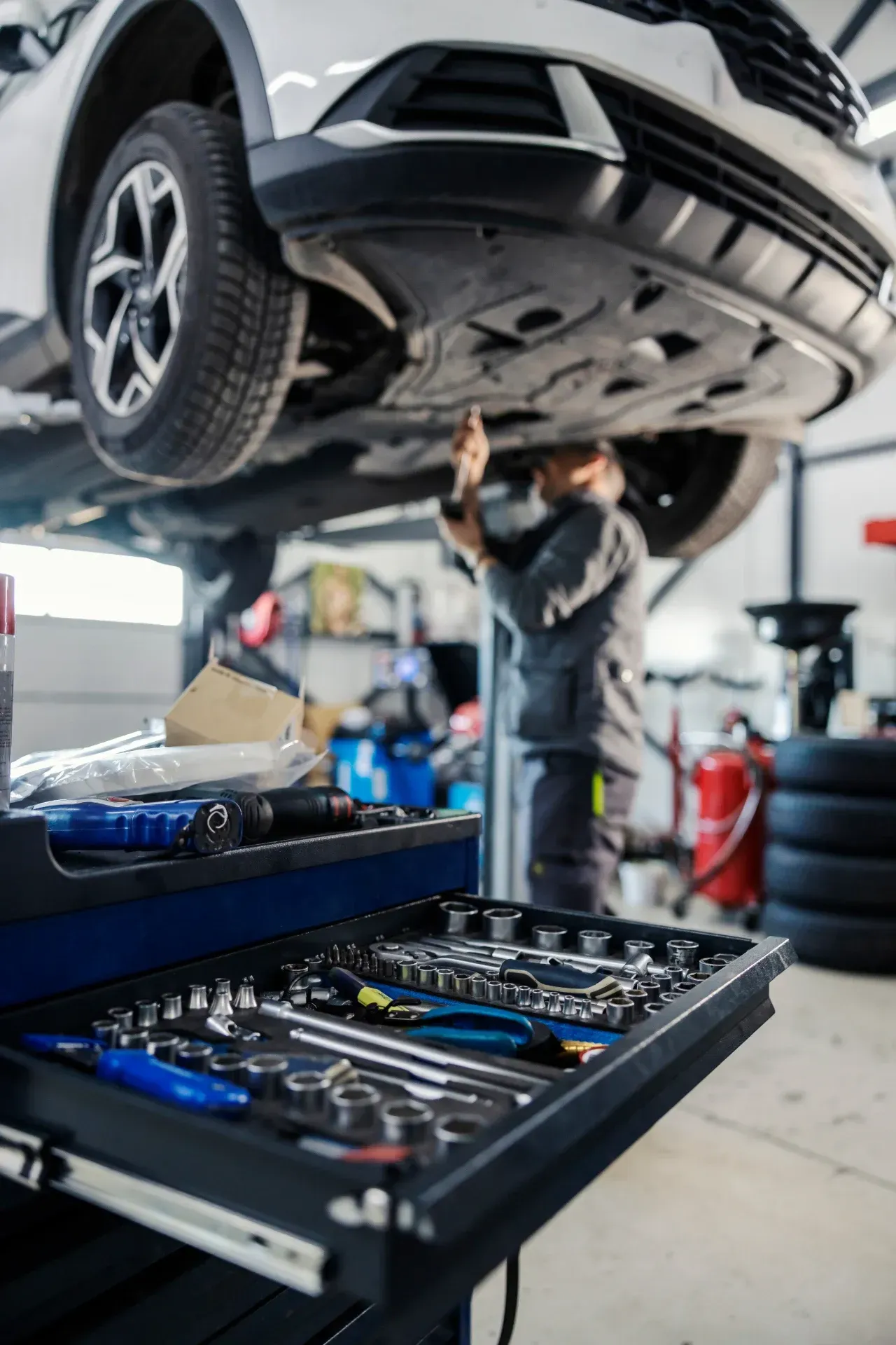 Ein Mechaniker arbeitet in einer Autowerkstatt unter einem angehobenen Auto; Werkzeuge befinden sich in einem nahegelegenen Tablett.