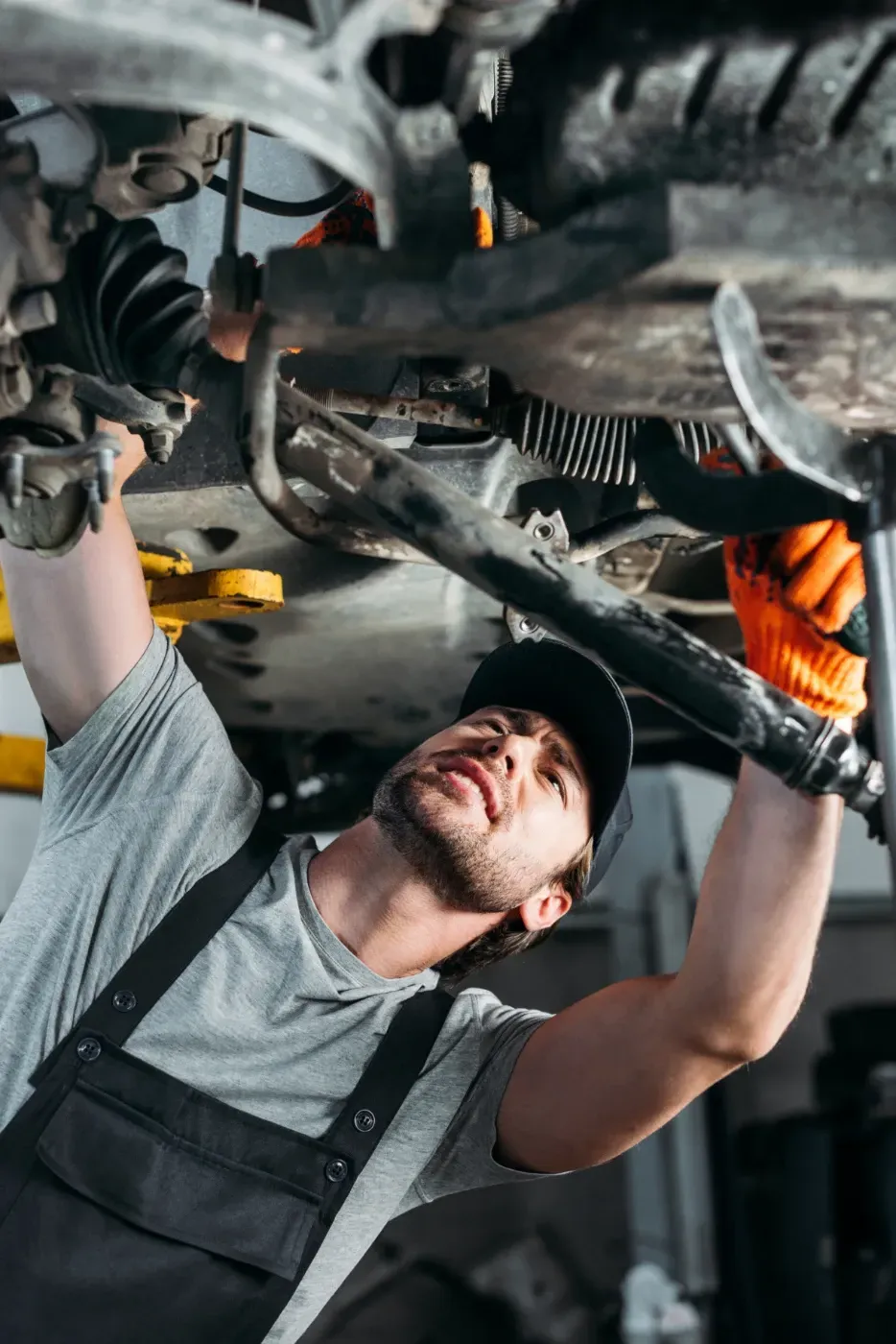Ein Mechaniker arbeitet unter einem Auto und überprüft mit orangefarbenen Handschuhen und einem Schraubenschlüssel die Aufhängung.