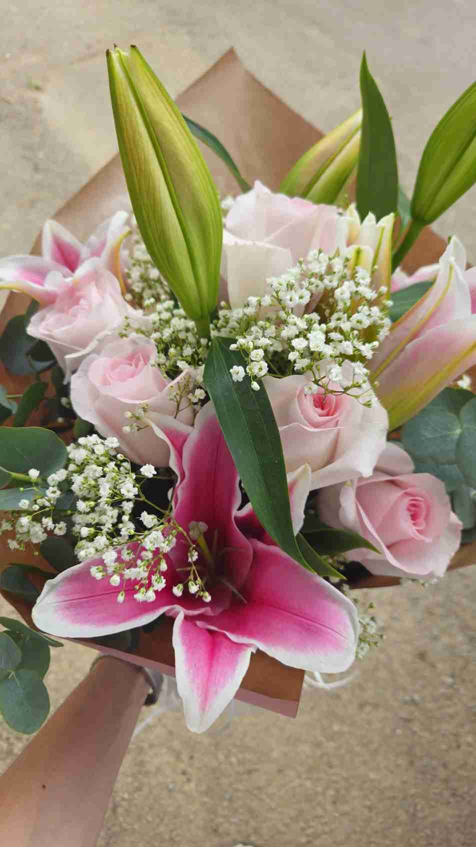 Ramo de rosas y lirios rosados, con giardineras y hojas verdes, envuelto en papel marrón.