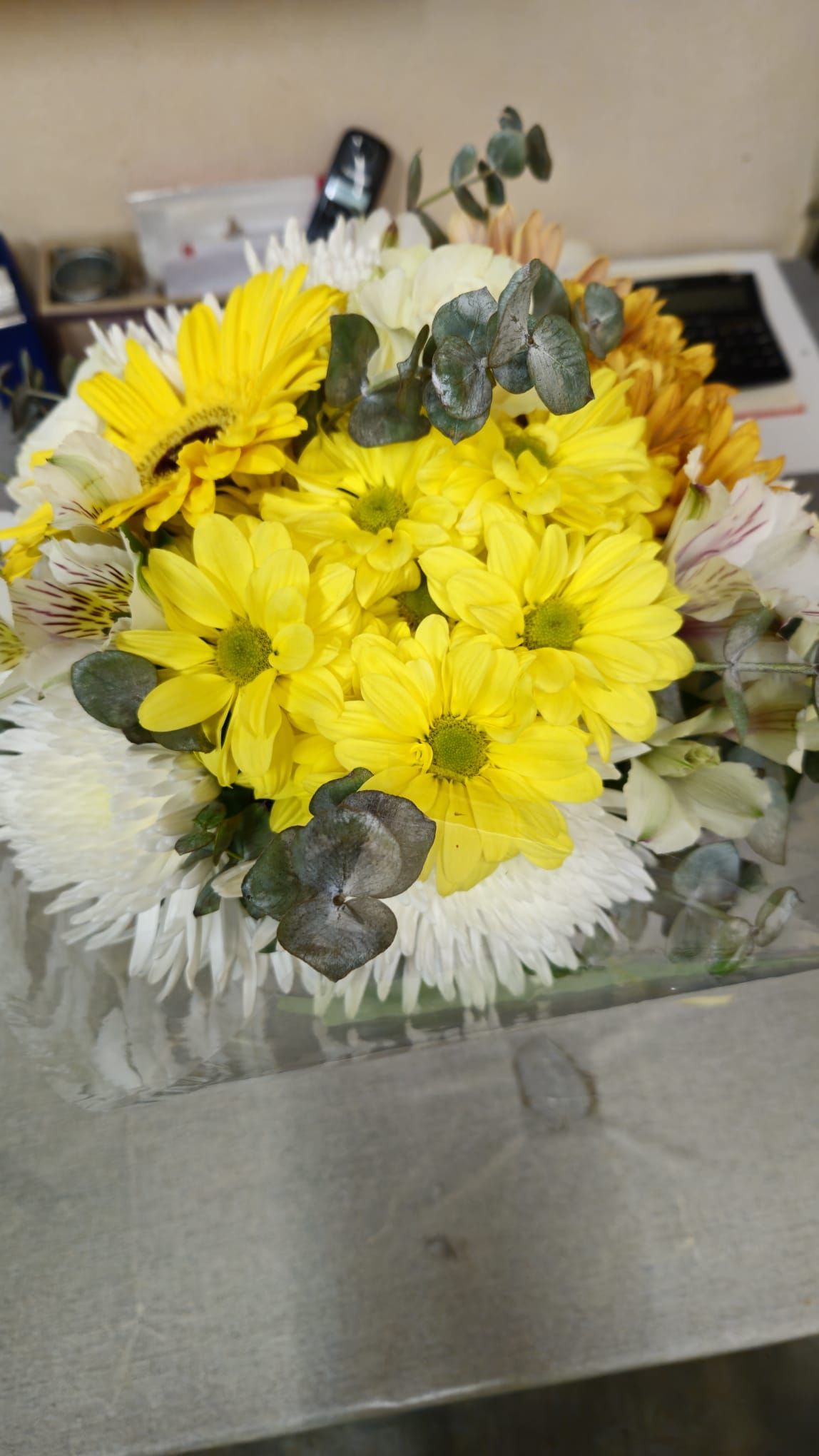 Ramo de flores amarillas y blancas con follaje verde sobre una superficie transparente.