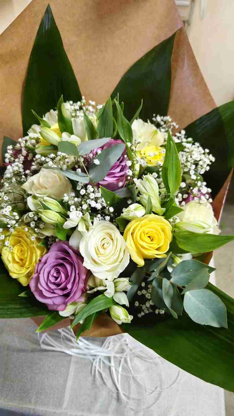 Ramo de rosas amarillas, blancas y moradas con vegetación, envuelto en papel marrón.