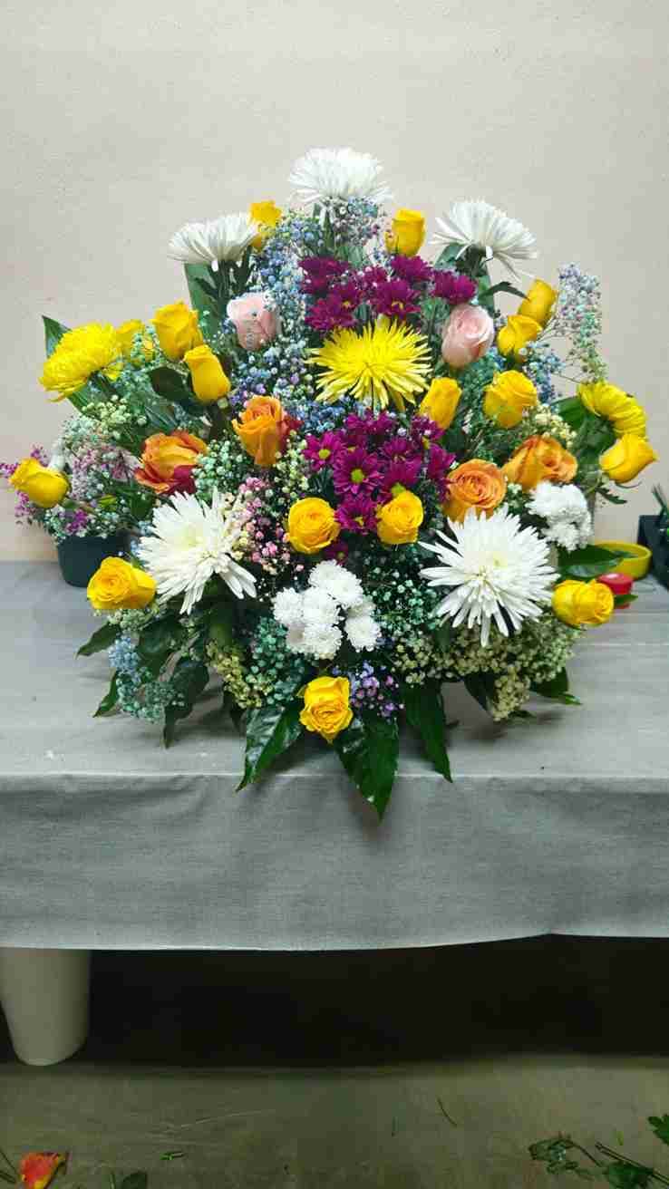 Arreglo floral colorido sobre una superficie gris; incluye flores amarillas, blancas, rosas, naranjas y violetas.