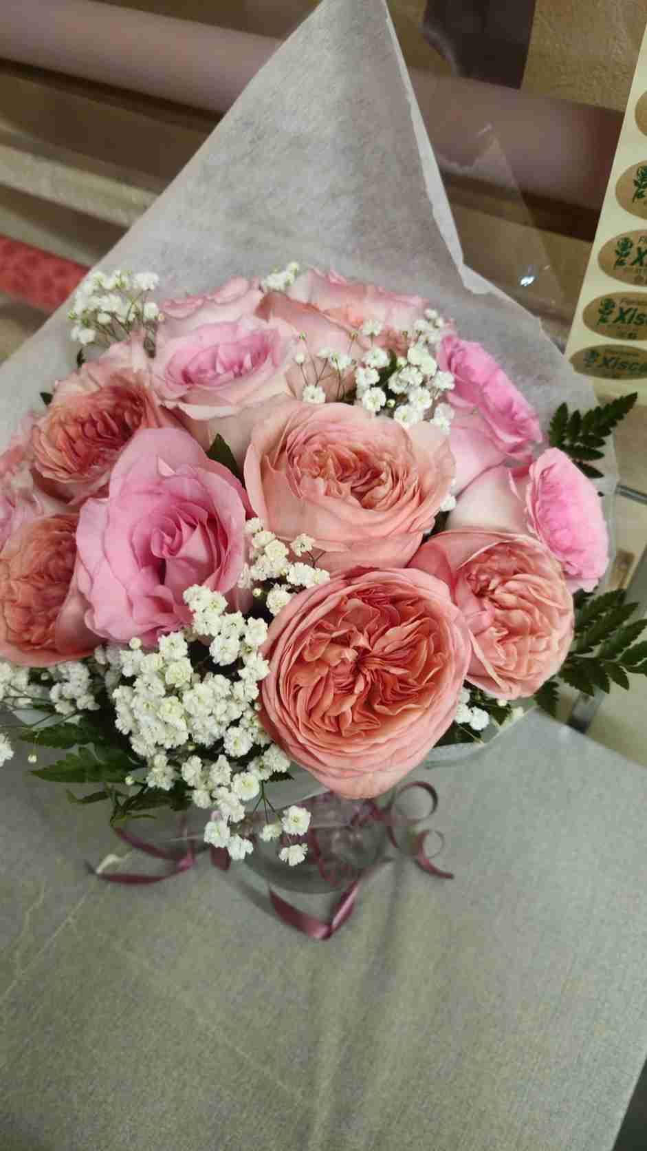 Ramo de rosas rosas y color melocotón, gipsófilas y vegetación envuelto en papel gris, atado con una cinta.