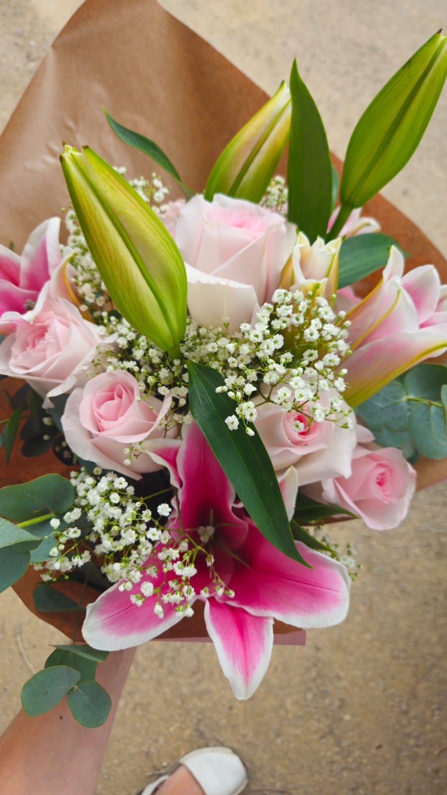 Ramo de rosas rosas y lirios con flores de baby's breath, envuelto en papel marrón, sostenido por una persona.