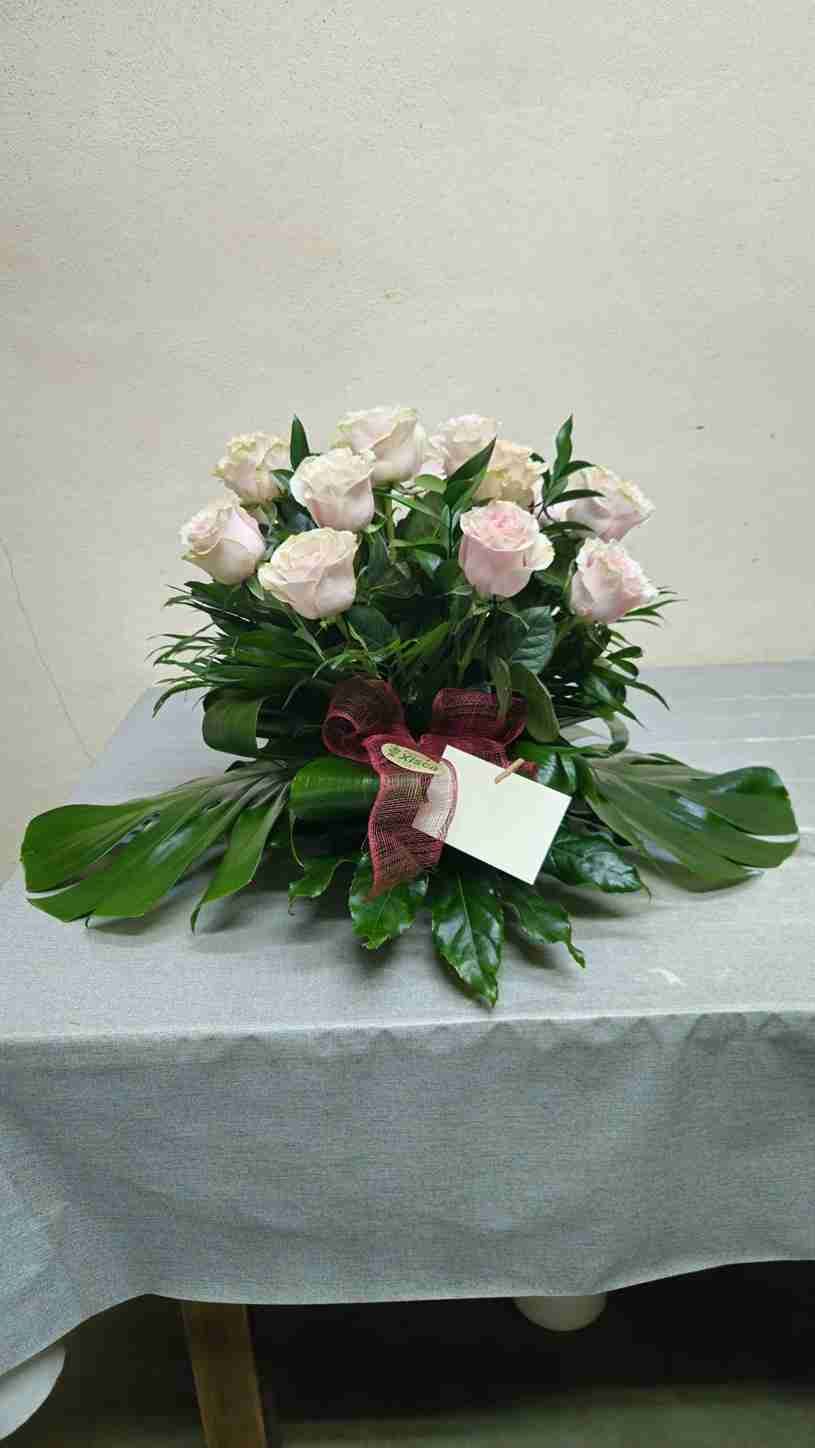 Un arreglo floral de rosas de color rosa claro y hojas verdes sobre una superficie gris, con una cinta decorativa y una tarjeta en blanco.