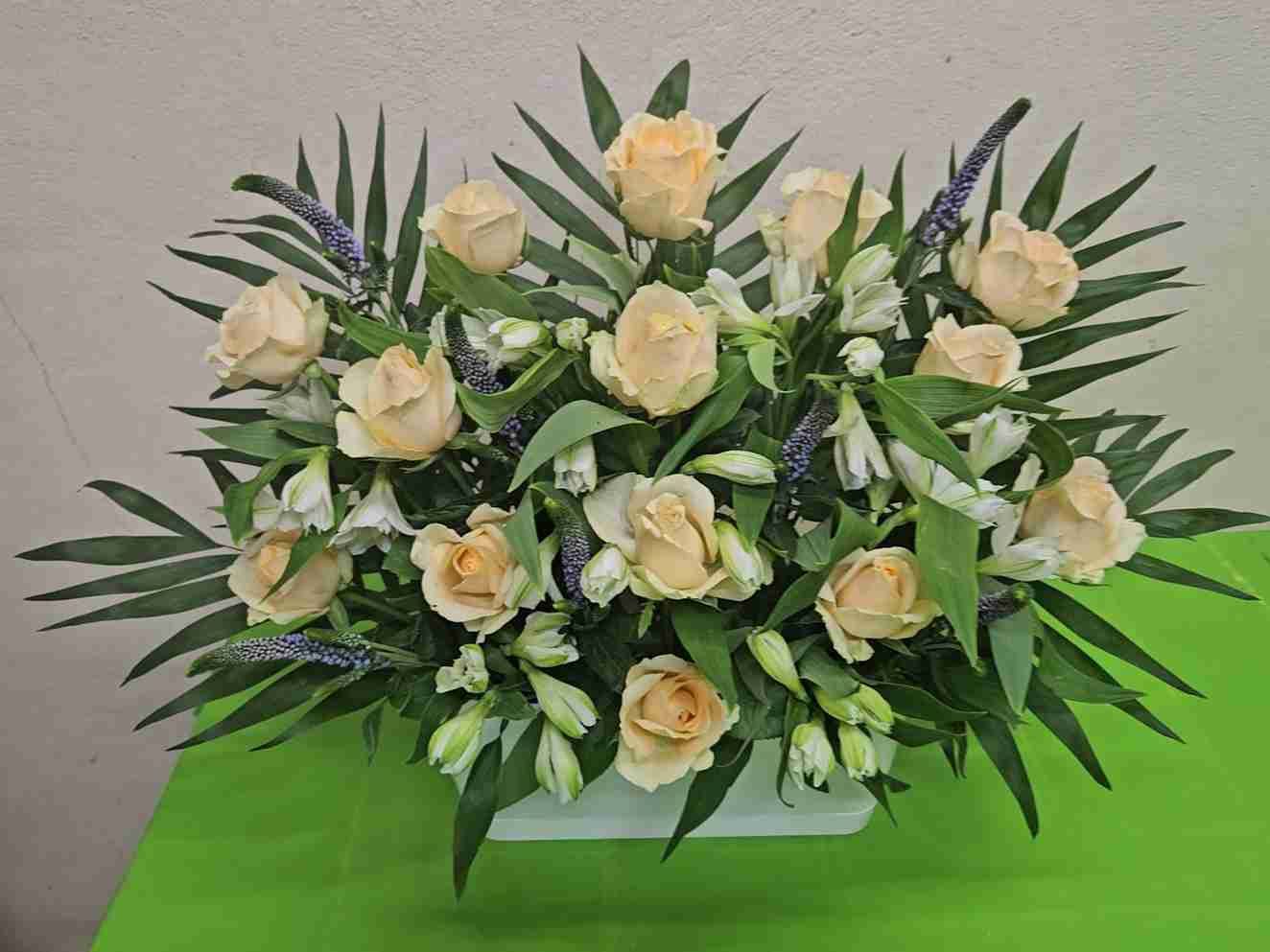 Arreglo floral en un recipiente cuadrado blanco, con rosas color durazno, flores blancas y vegetación sobre una superficie verde.