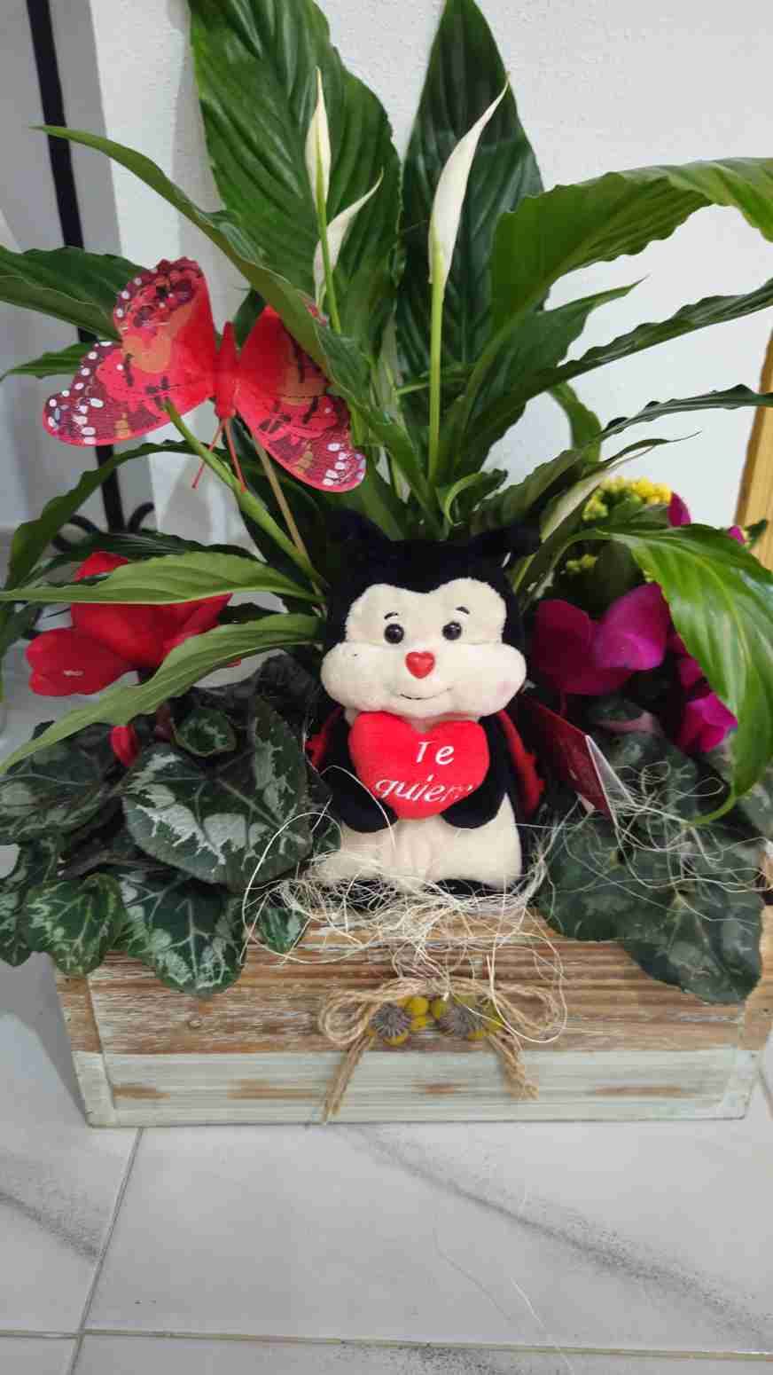 Un arreglo floral en una caja de madera con una mariquita de peluche sosteniendo un corazón que dice