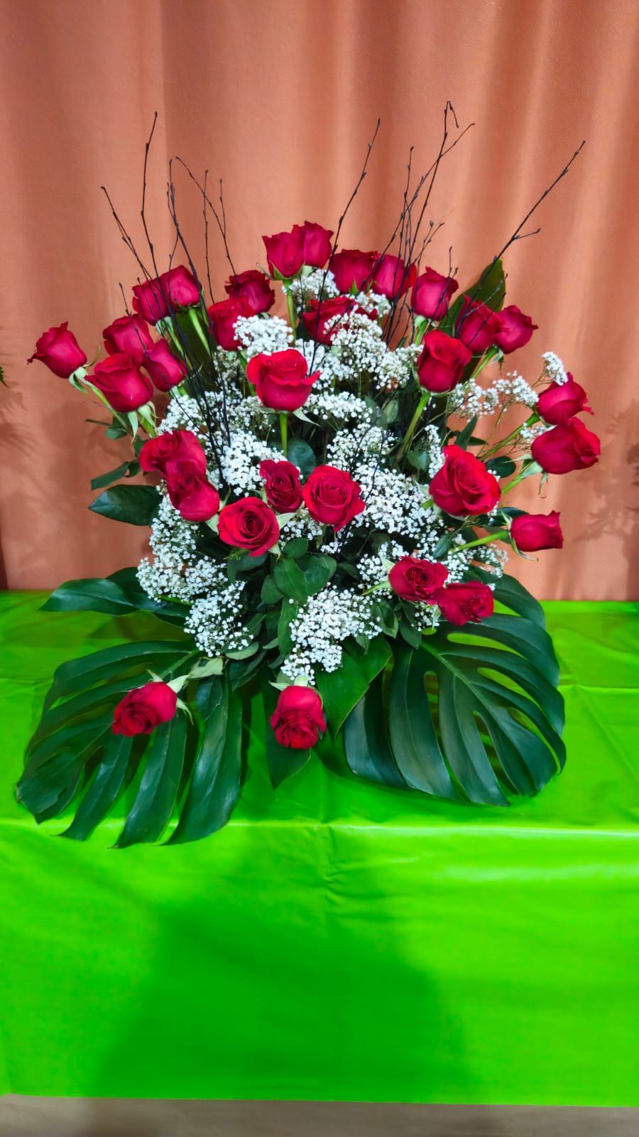 Arreglo floral de rosas rojas y gitanos sobre superficie verde, con grandes hojas verdes y fondo color melocotón.