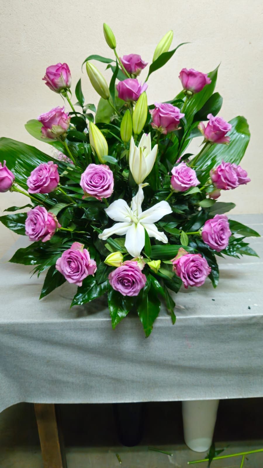 Rosas moradas y lirios blancos en un arreglo floral sobre una mesa. Hojas verdes como fondo.