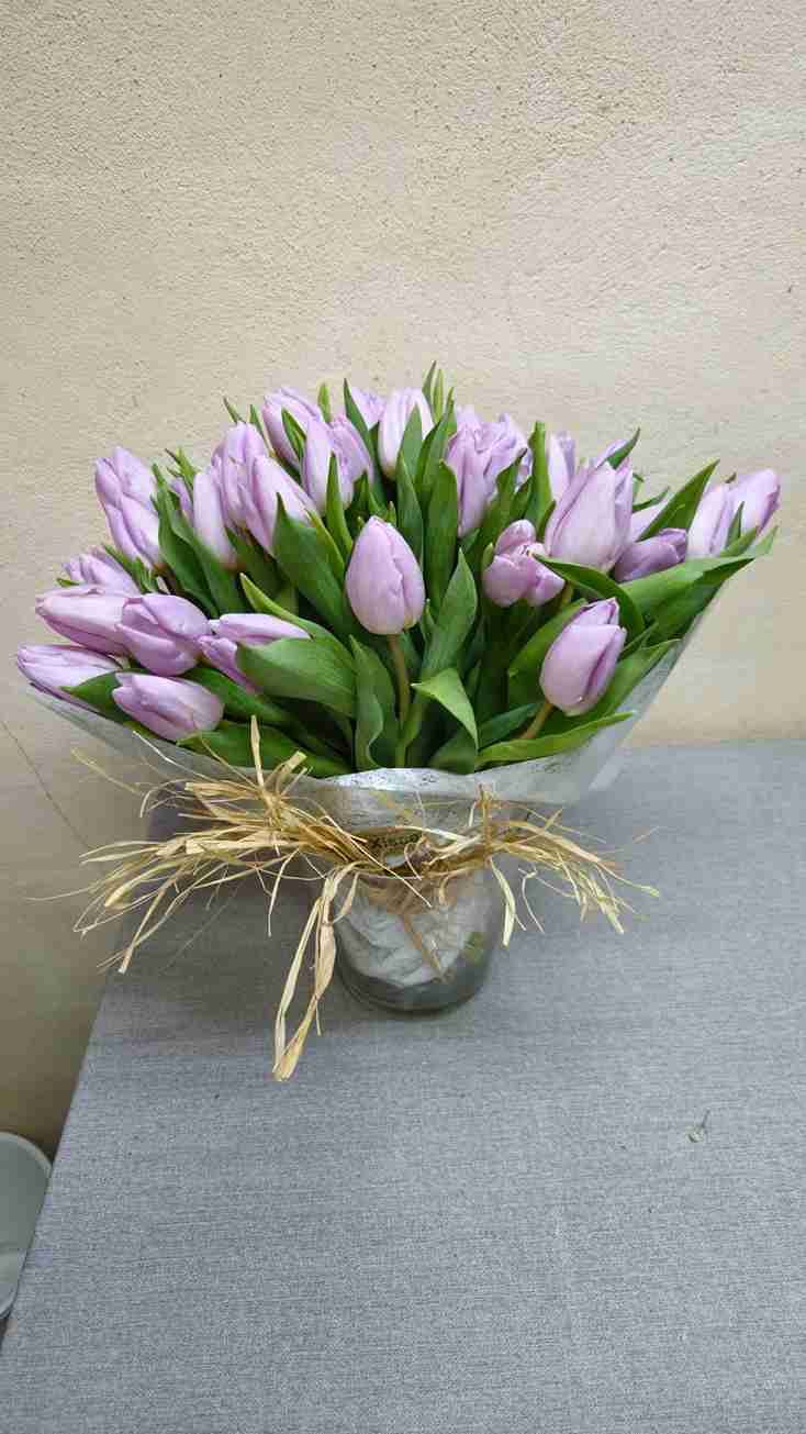 Ramo de tulipanes lavanda en jarrón de cristal, atado con rafia, sobre una superficie gris.