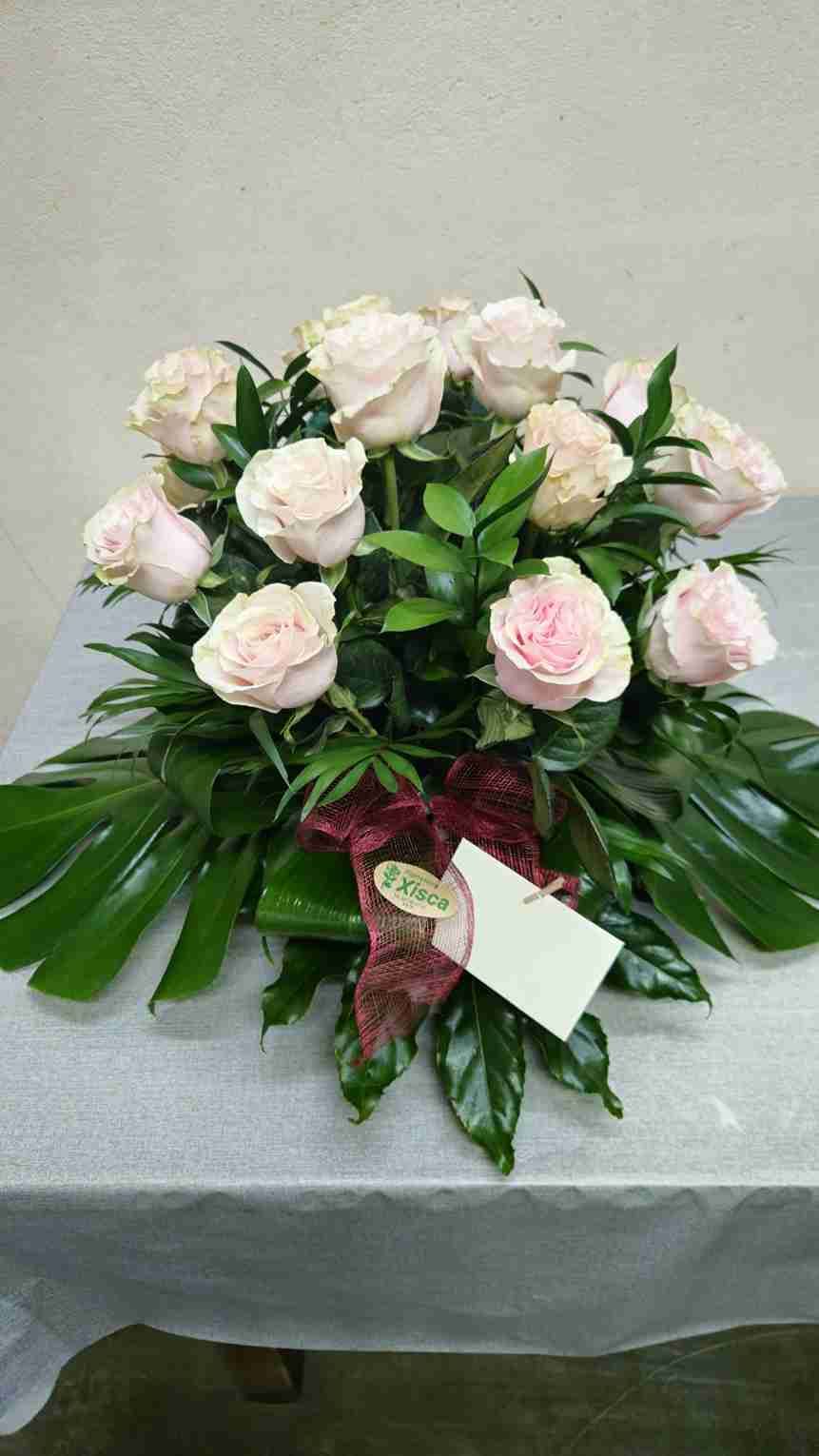 Rosas de color rosa pálido en un arreglo con hojas verdes, un lazo rojo y una tarjeta en blanco sobre una mesa.