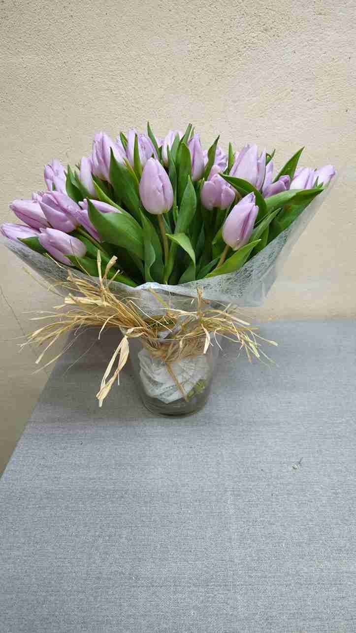 Tulipanes morados en un jarrón de cristal, envueltos en papel decorativo, sobre una superficie gris.