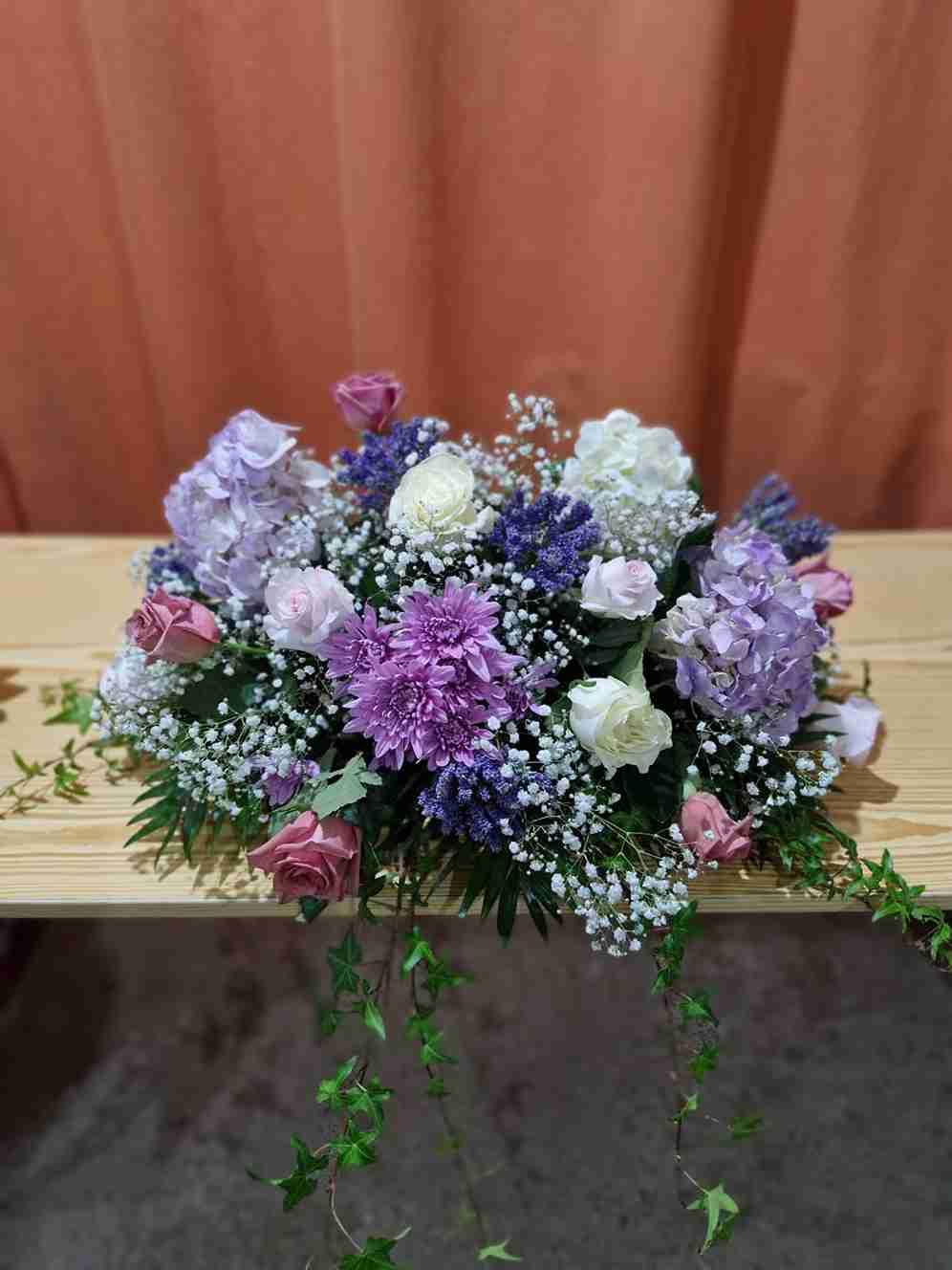 Arreglo floral con flores de color violeta, rosa y blanco sobre una superficie de madera con hiedra.