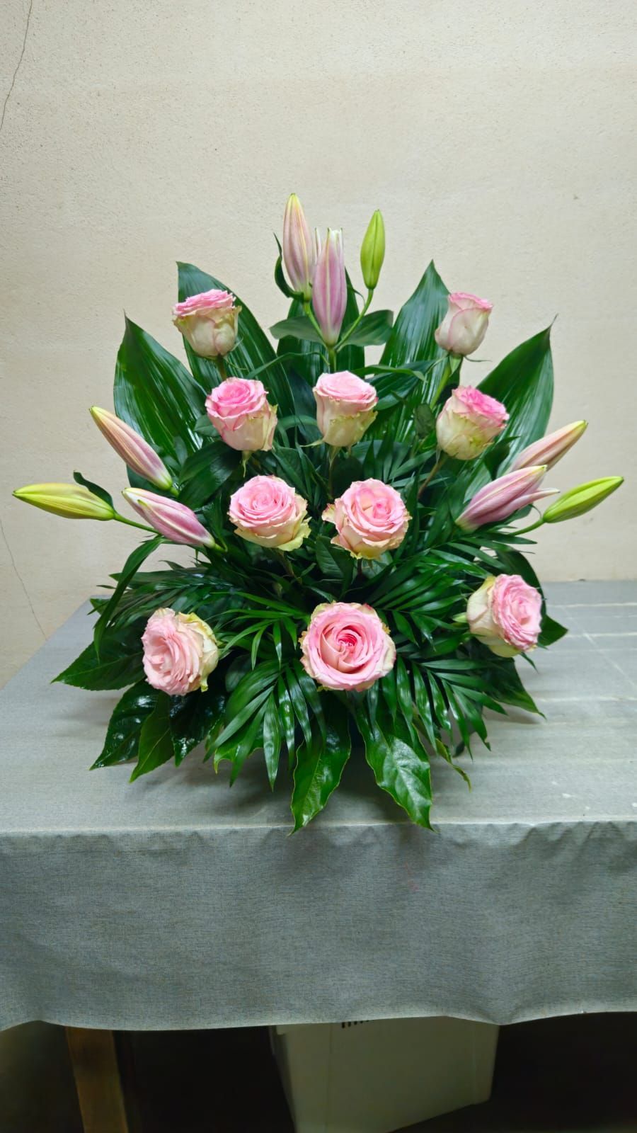 Arreglo floral de rosas rosadas y lirios, rodeados de hojas verdes sobre una superficie gris.