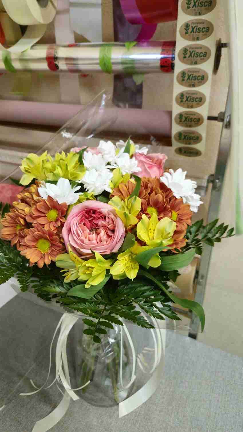 Ramo de flores mixtas en un jarrón de vidrio; incluye flores naranjas, blancas, rosas y amarillas con vegetación, cinta atada alrededor del jarrón.